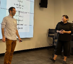 a smiling woman dressed in black is turned to her left facing a smiling man in a white button-down shirt with tan pants